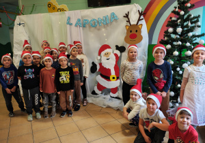 Przedszkolaki w czapkach Mikołaja stoją na tle dekoracji Laponii.