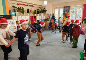 Przedszkolaki w czapkach Mikołaja stoją w parach na dywanie.