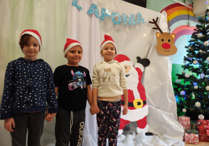 Dwie dziewczynki i jeden chłopiec stoją w czapkach Mikołaja na tle dekoracji Laponii.