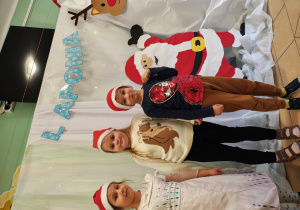 Dwie dziewczynki i jeden chłopiec stoją w czapkach Mikołaja na tle dekoracji Laponii.