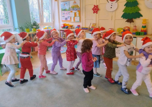Przedszkolaki z grupy I, w czapkach Mikołaja stoją w wężyku na dywanie.