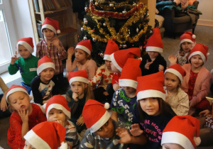 Przedszkolaki z grupy III w czapkach Mikołaja siedzą na dywanie. W tle choinka.