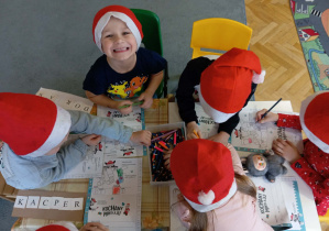 Przedszkolaki w czapkach Mikołaja kolorują świąteczne obrazki.