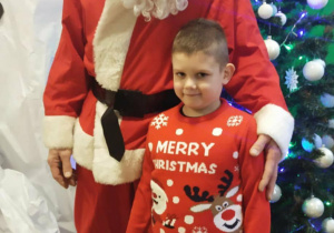 Chłopiec w świątecznym sweterku stoi z Mikołajem na tle choinki.