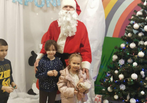 Przedszkolaki stoją z Mikołajem obok choinki.