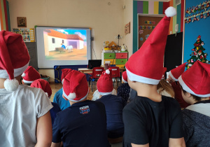 Przedszkolaki w czapkach Mikołaja oglądają film animowany.