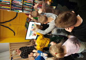 Przedszkolaki stoją z uniesionymi ramionami przed ekranem komputera. Kobieta w sukience w kratę stoi obok ekranu.