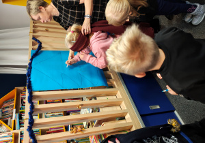 Dziewczynka z zasłoniętymi oczami z pomocą kobiety w sukience w kratkę,rysuje bombkę na kartonie z choinką.