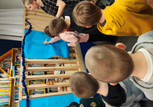 Dziewczynka z zasłoniętymi oczami z pomocą kobiety w sukience w kratkę,rysuje bombkę na kartonie z choinką.