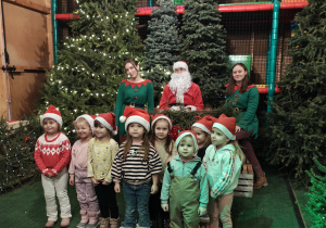 Przedszkolaki w czapkach Mikołaja stoją przed saniami Mikołaja. W saniach siedzi Mikołaj. Obok sań stoja dwie kobiety przebrane za elfy.