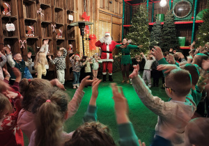Przedszkolaki wraz z Mikołajem i kobietami przebranymi za elfy stoją w kole z uniesionymi rękami.