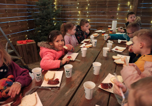 Przedszkolaki siedzą przy drewnianych stołach i jedzą kiełbaski.