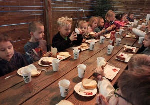 Przedszkolaki siedzą przy drewnianych stołach i jedzą kiełbaski.