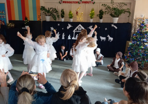 Dziewczynki w białych sukienkach tańczą na dywanie. W tle biała świąteczna dekoracja.