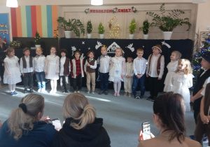 Przedszkolaki w przebraniach pastuszków, trzech kroli i aniołków stoją w półkolu na dywanie. W tle świąteczna dekoracja.