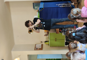 Mężczyzna gra na trąbce. Dzieci siedzą przed nimi na dywanie.