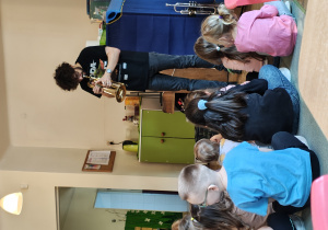 Mężczyzna gra na trąbce. Dzieci siedzą przed nimi na dywanie.