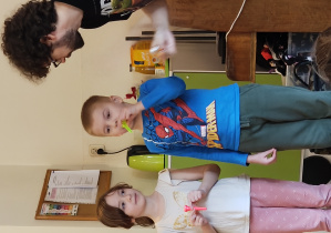 Chłopiec dmucha w ustnik. Dziewczynka trzyma ustnik. Obok stoi mężczyzna.