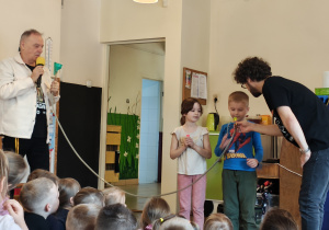 Chłopiec dmucha w ustnik podłączony do rurki. Dziewczynka trzyma ustnik. Obok stoi mężczyzna trzymający ustnik. Drugi mężczyzna trzyma końcówkę instrumentu.