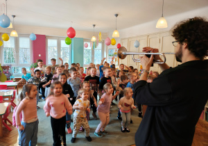 Mężczyzna gra na trąbce. Dzieci stoją przed nim na dywanie.