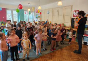 Mężczyzna mówi do mikrofonu. Dzieci stoją przed nimi na dywanie.