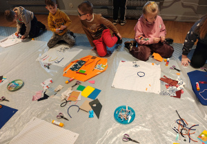 Przedszkolaki siedzą na podłodze. Przed nimi kwadratowe kawałki materiału, na których dzieci tworzą obrazki ze sznurków, materiału, papieru.