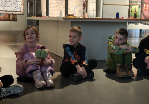 Dzieci siedzą na podłodze w kole. Dziewczynka trzyma pluszową opuncję figową.