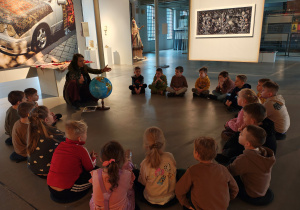 Kobieta z globusem siedzi na podłodze muzeum. Dzieci siedzą w kole.