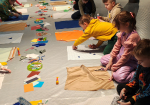 Przedszkolaki siedzą na podłodze. Przed nimi kwadratowe kawałki materiału, na których dzieci tworzą obrazki ze sznurków, materiału, papieru.