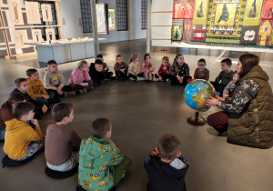 Kobieta z globusem siedzi na podłodze muzeum. Dzieci siedzą w kole.