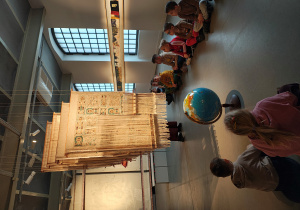 Na środku wiszą materiały. Przed nimi stoi globus. Dzieci siedzą po dwóch stronach globusa.