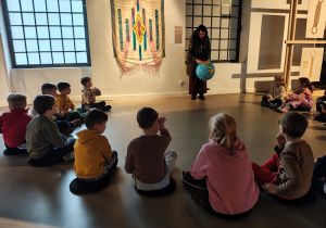 Kobieta stoi trzymając globus. Dzieci siedzą w półkolu przed nią. Na ścianie za kobietą wisi materiał.