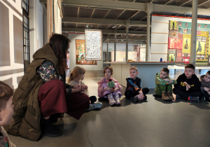 Kobieta wraz z dziećmi siedzi na podłodze w kole. Dziewczynka trzyma pluszową opuncję figową.