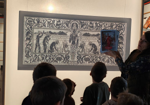 Przedszkolaki stoją przed obrazem. Obok obrazu stoi kobieta, która trzyma zdjęcia. .