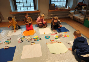 Przedszkolaki siedzą na podłodze. Przed nimi kwadratowe kawałki materiału, na których dzieci tworzą obrazki ze sznurków, materiału, papieru.