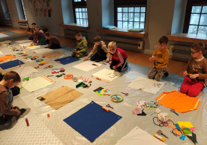 Przedszkolaki siedzą na podłodze. Przed nimi kwadratowe kawałki materiału, na których dzieci tworzą obrazki ze sznurków, materiału, papieru.