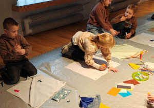 Przedszkolaki siedzą na podłodze. Przed nimi kwadratowe kawałki materiału, na których dzieci tworzą obrazki ze sznurków, materiału, papieru.