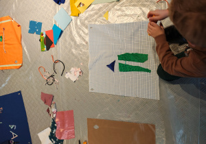 Chłopiec siedzi na podłodze, przed nim leży materiał. Chłopiec tworzy obrazek ze sznurków, materiału, papieru.