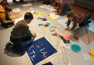 Przedszkolaki siedzą na podłodze. Przed nimi kwadratowe kawałki materiału, na których dzieci tworzą obrazki ze sznurków, materiału, papieru.