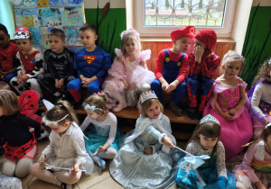 Przebrane za bohaterów bajek oraz inne postacie przedszkolaki siedzą na podłodze i na ławce.