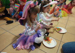 Dwie dziewczynki siedzą na podłodze i nawijają zieloną włóczkę na widelec. W tle przedszkolaki siedzace na podłodze.