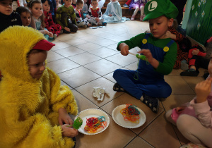 Dwóch chłopców siedzi na podłodze i nawija zieloną włóczkę na widelec. W tle przedszkolaki siedzace na podłodze.
