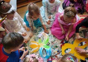 Na środku leży pokolorowana, papierowa maska. Dzieci siedzą na ceracie i ozdabiają maskę.