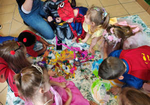 Na środku leży pokolorowana, papierowa maska. Dzieci siedzą na ceracie i ozdabiają maskę.