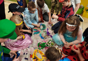 Na środku leży pokolorowana, papierowa maska. Dzieci siedzą na ceracie i ozdabiają maskę.