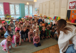 Przedszkolaki kucają na dywanie. Kobieta z mikrofonem kuca przed nimi.