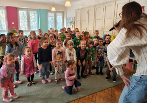 Przedszkolaki stoją na dywanie. Kobieta z mikrofonem stoi przed nimi.