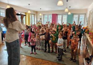 Przedszkolaki z wyciagniętymi w górę ramionami stoją na dywanie. Kobieta z mikrofonem stoi przed nimi z wyciagniętą ku górze jedną ręką..