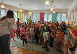 Przedszkolaki stoją na dywanie. Kobieta z mikrofonem stoi przed nimi.