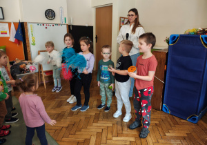Trzy diewczynki stoją z pomponami. Trzech chłopców stoi z instrumentami. Za nimi stoi kobieta z mikrofonem. Przed nimi stoją przedszkolaki.